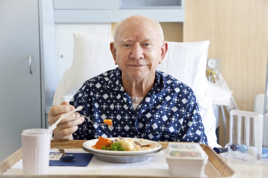 An elderly man sits in a hospital bed, wearing a hospital gown. He is eating from a hospital tray which includes a healthy meal and a drink.