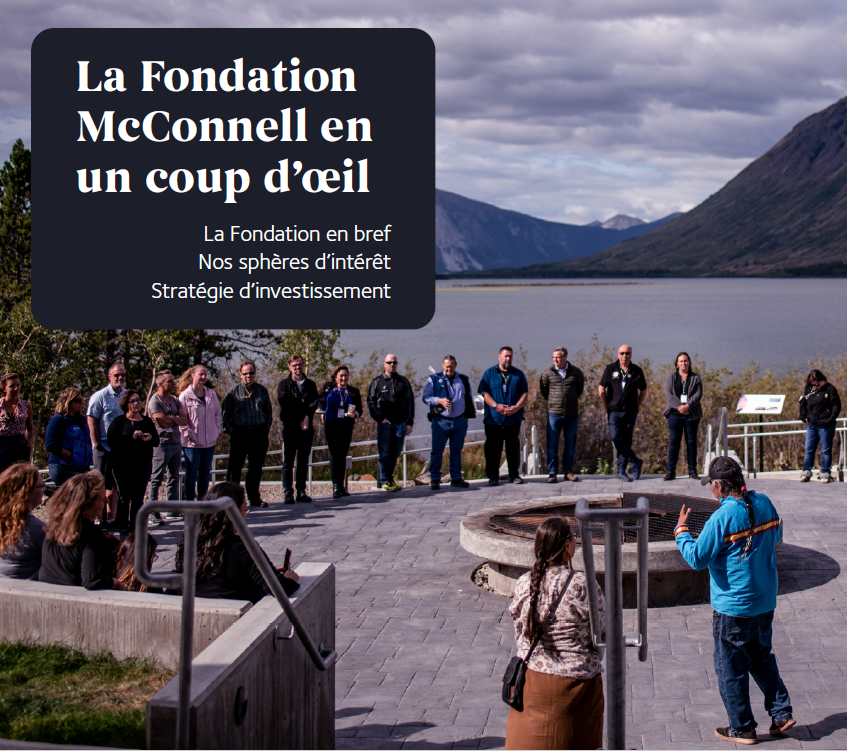 At the border of a lake surrounded by mountains, a large group of individuals stand in a circle around a fire pit. An Indigenous man with a long braid and a woman address them. This meeting happened at the Summer Institute, at the Kwanlin Dün Cultural Centre, Yukon in 2019.