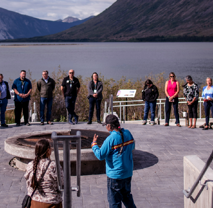 At the border of a lake surrounded by mountains, a large group of individuals stand in a circle around a fire pit. An Indigenous man with a long braid and a woman address them. This meeting happened at the Summer Institute, at the Kwanlin Dün Cultural Centre, Yukon in 2019.