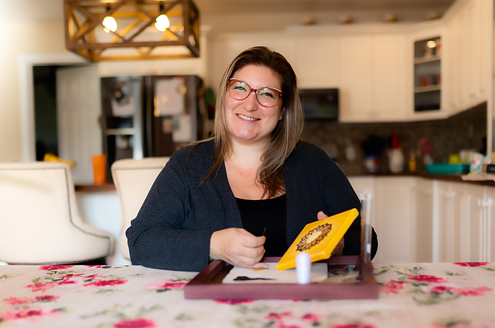 Candace Kahnekanonha Snook looks at the camera holding a needle and beading artwork, wearing glasses and smiling.