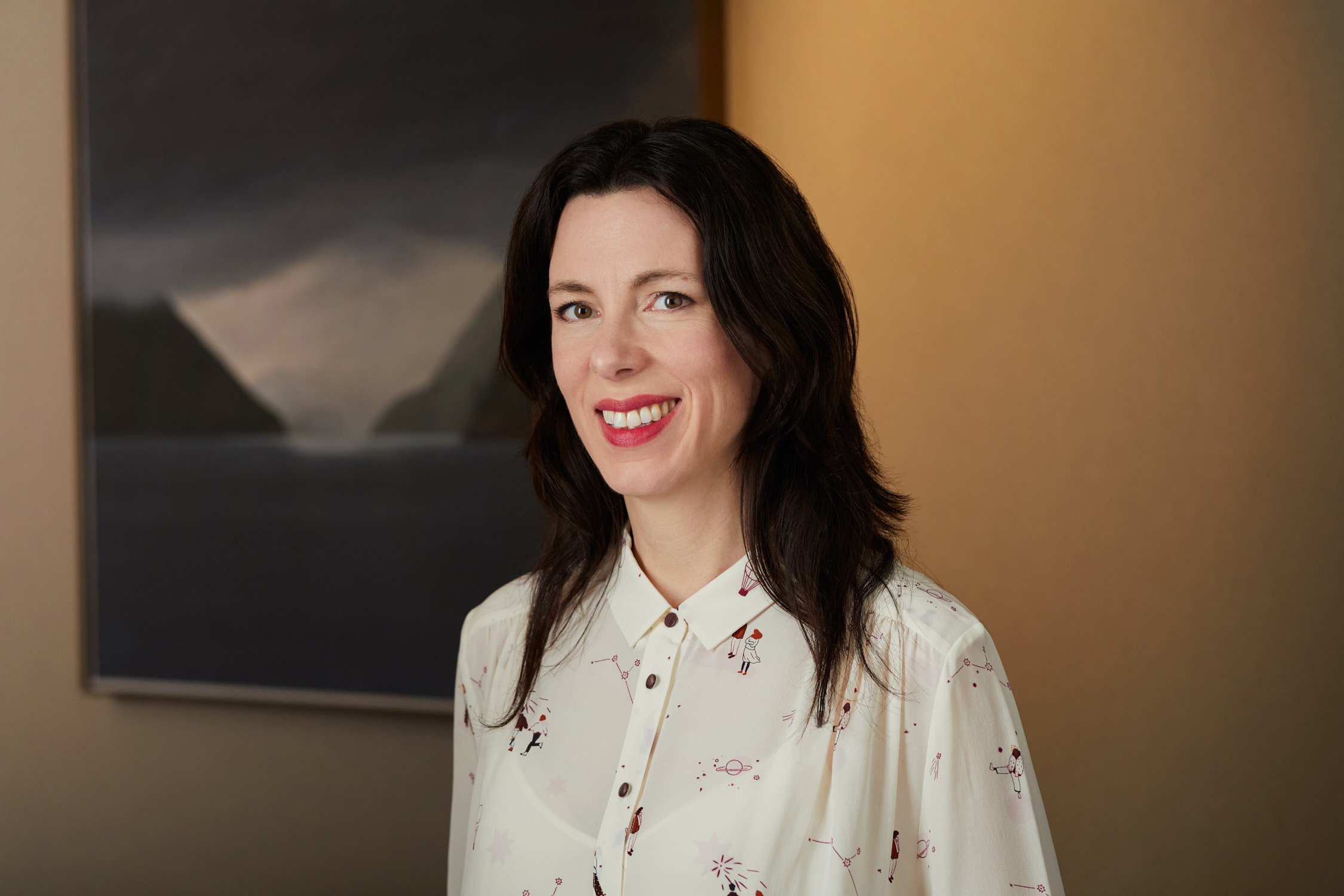 Photo de Maggie MacDonald, avec une œuvre d'art à l'arrière-plan représentant un lac, avec des montagnes nuageuses dans les gris et les bleus sombres.