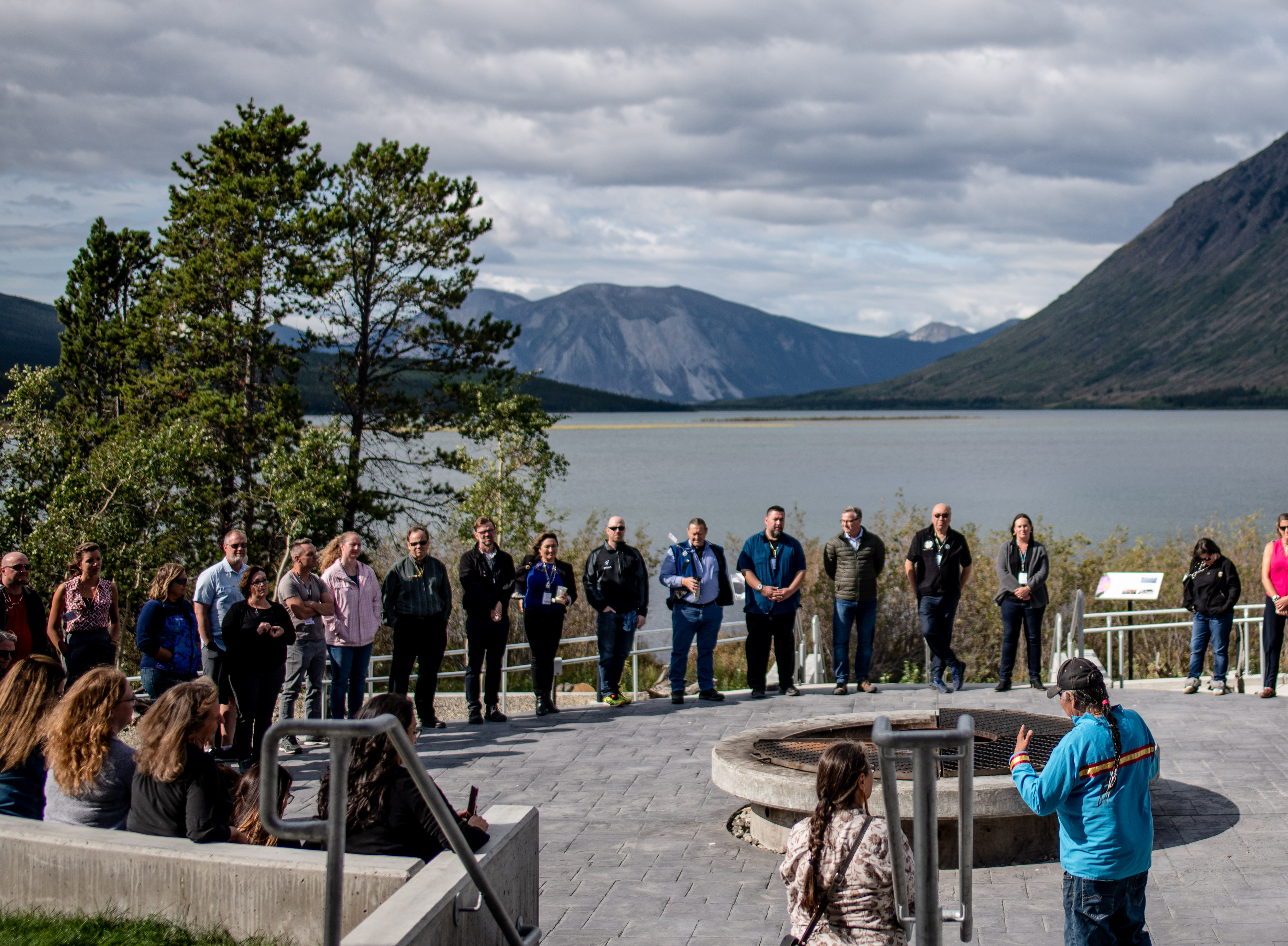 At the border of a lake surrounded by mountains, a large group of individuals stand in a circle around a fire pit. An Indigenous man with a long braid and a woman address them.
